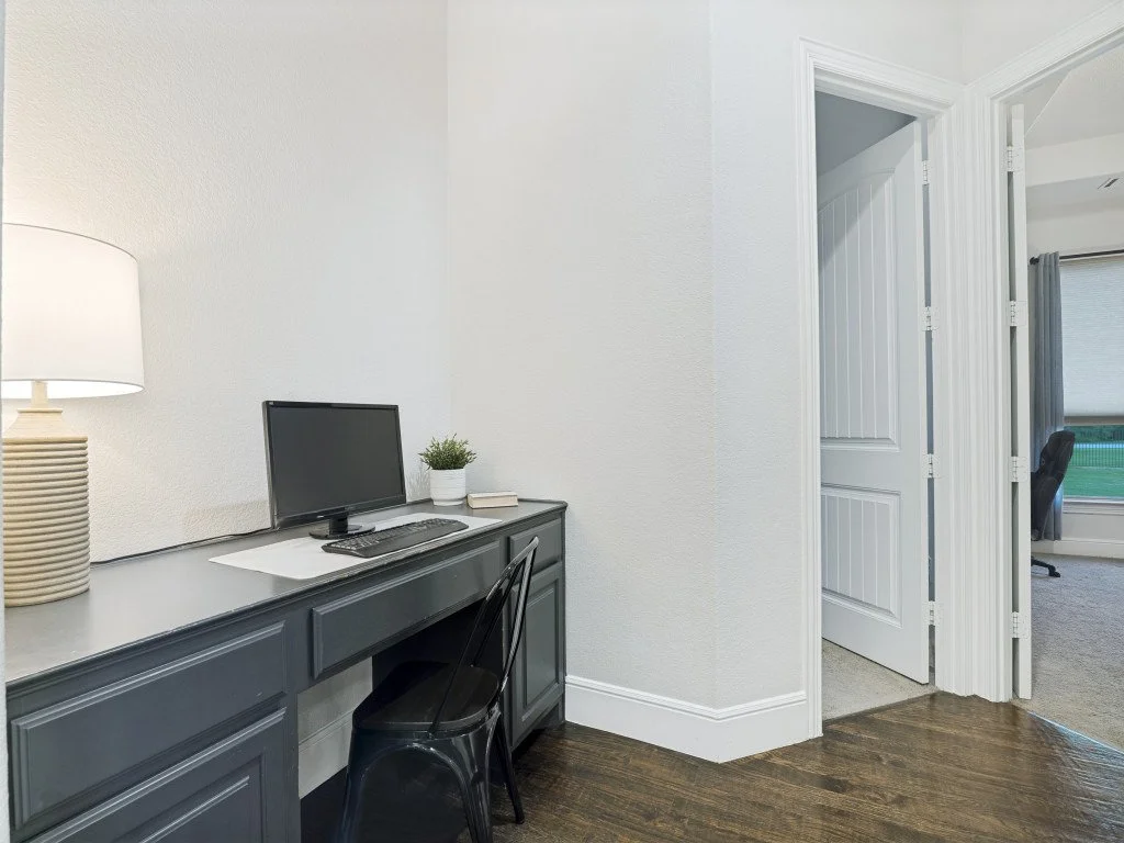 Home office area with a gray desk, a computer monitor, a lamp, a potted plant, and a door leading to an adjacent room with a window.