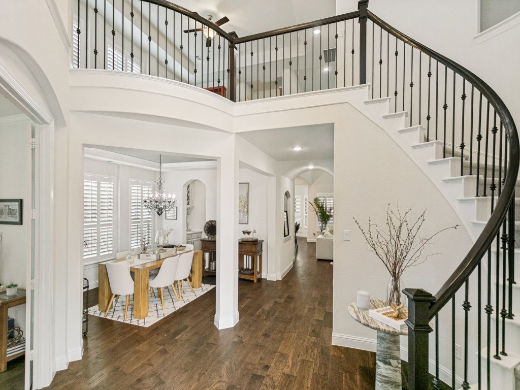 Interior view of a house entryway featuring a stairwell with black railing, a dark wood floor, and a dining area with a chandelier, white chairs, and large windows with shutters.