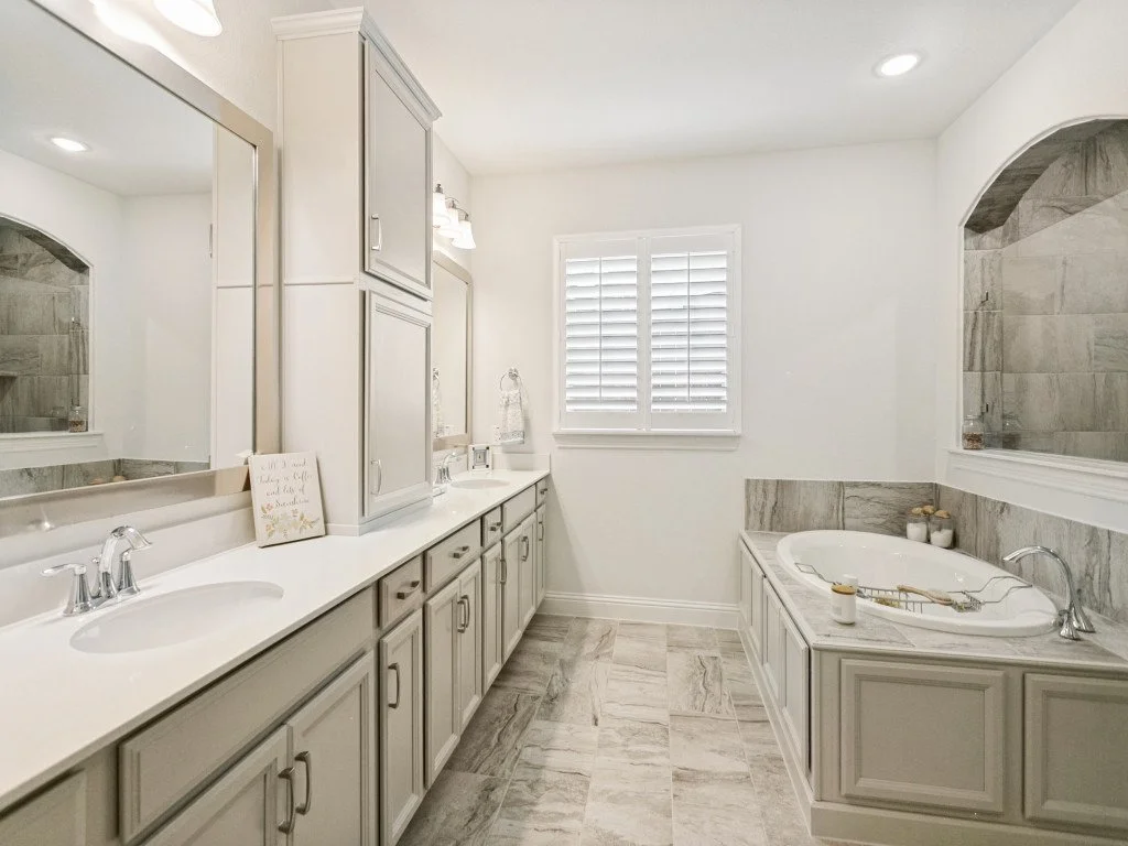 Bright bathroom with dual sinks, light gray cabinets, and a large mirror. On the right, a built-in bathtub with stone tiles surrounding it. A window with plantation shutters is centered on the back wall. Recessed ceiling lights. The floor and wall ti