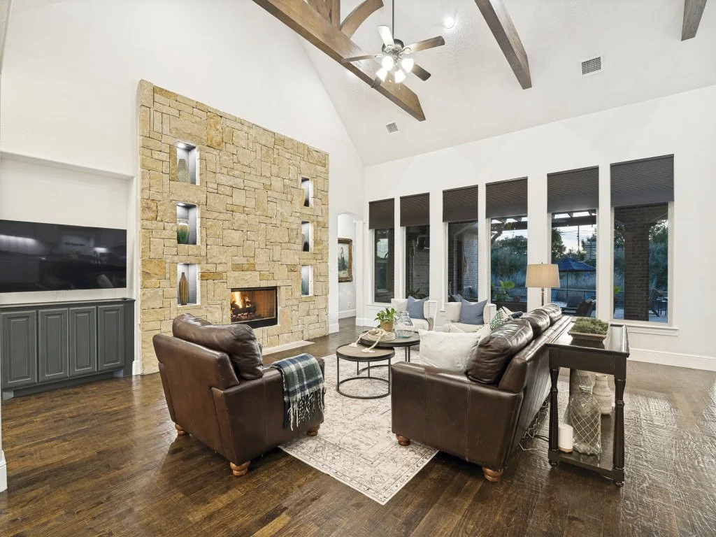 Living room with a stone fireplace, leather chairs, a sofa, a ceiling fan with lights, hardwood floors, and large windows.