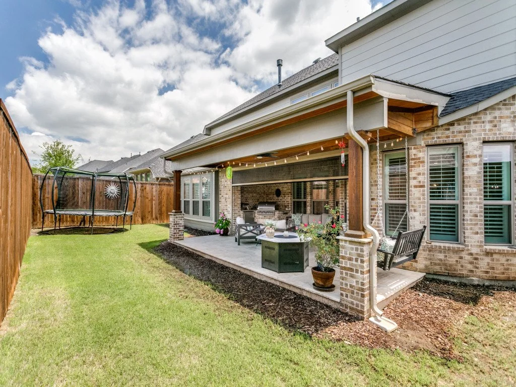 Backyard with a covered patio, outdoor furniture, plants, and a trampoline, enclosed by a wooden fence under a partly cloudy sky.