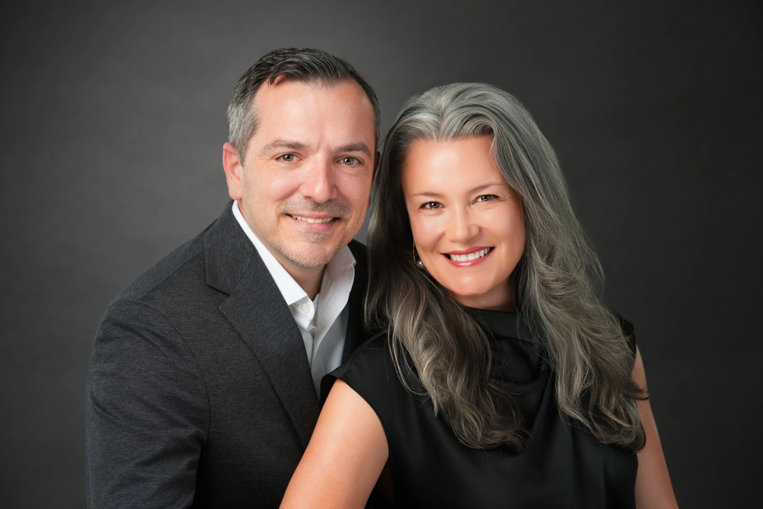 A middle-aged man with gray hair and a woman with gray hair styled in loose curls, both dressed in formal attire, smiling in a professional studio portrait with a dark background.