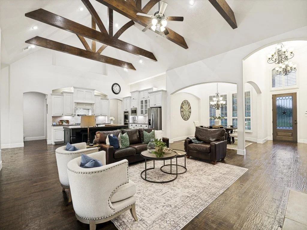 Open-concept living room and kitchen with white cabinets, vaulted ceiling with exposed wooden beams, ceiling fans, hardwood flooring, and decorative rugs. Furniture includes leather sofas, armchairs, and a coffee table. Large window and front door with glass panels are visible.