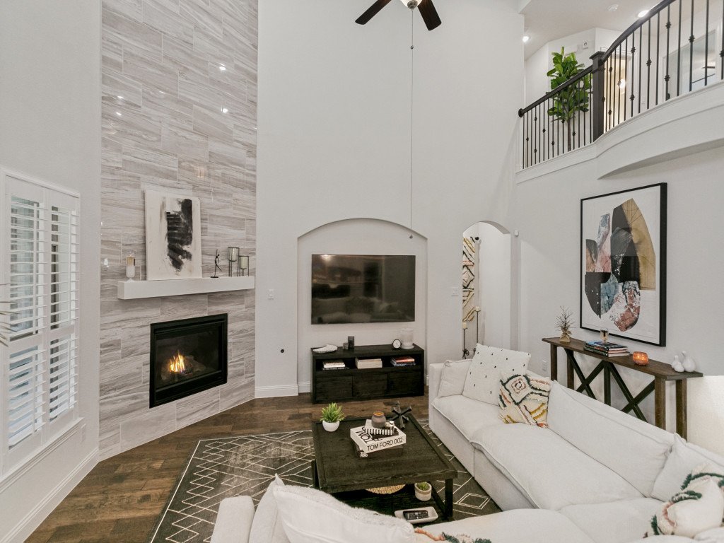 A living room with a fireplace, a mounted flat-screen TV, white sofas, a wooden coffee table, and wall art on a white wall. There's a ceiling fan and a second-floor balcony with a railing.