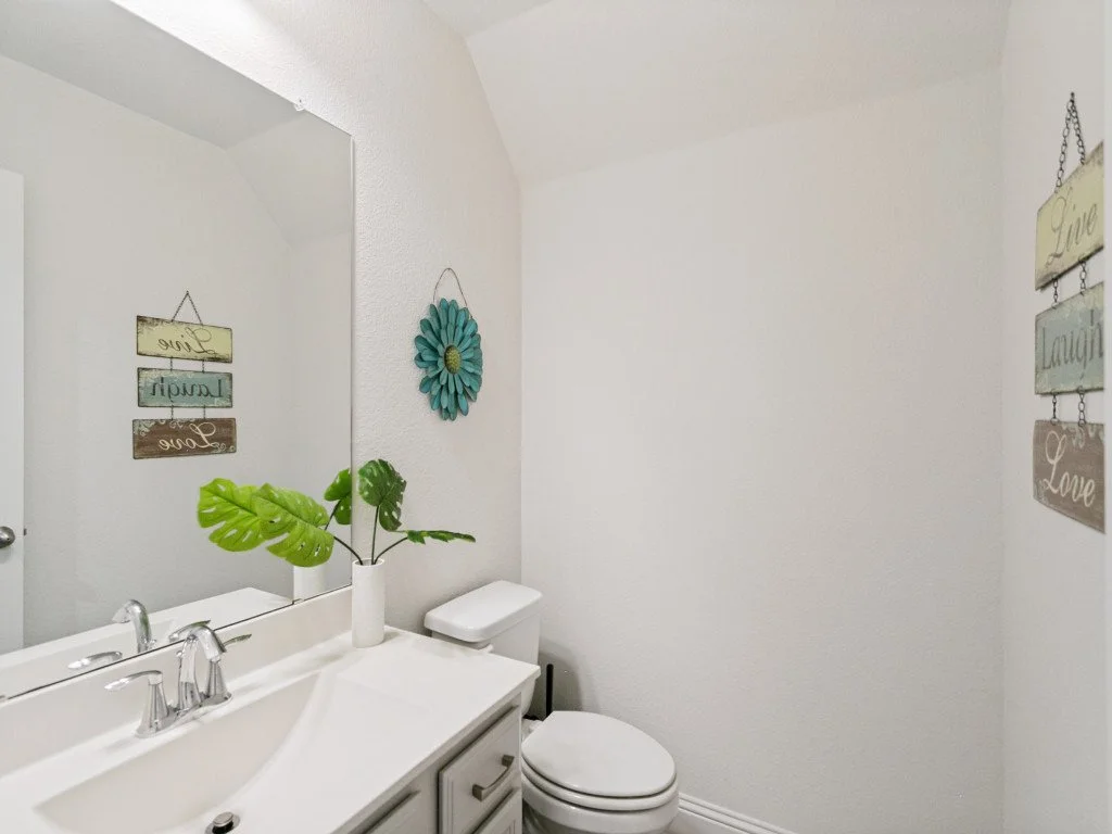 A small bathroom with white walls, a mirror, a sink with silver fixtures, a cabinet, a toilet, a potted plant with large green leaves, and decorative wall art with inspirational words.