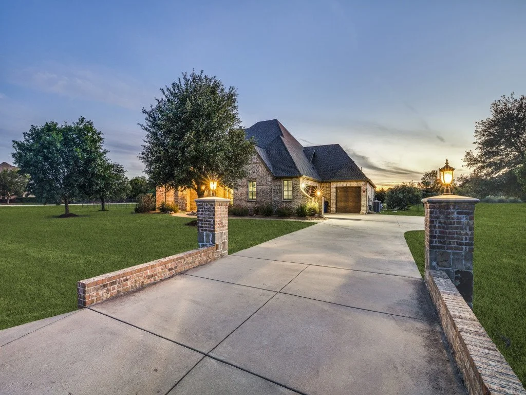 A brick house with a driveway, surrounded by a grassy lawn and trees, lit by outdoor lanterns at dusk.