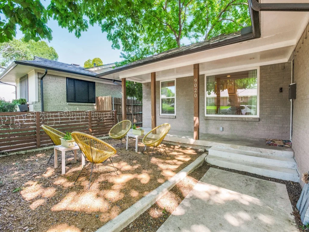A backyard patio with three yellow outdoor chairs, two white side tables with plants, a concrete porch with steps, and a wooden fence with string lights, surrounded by green trees and neighboring houses.