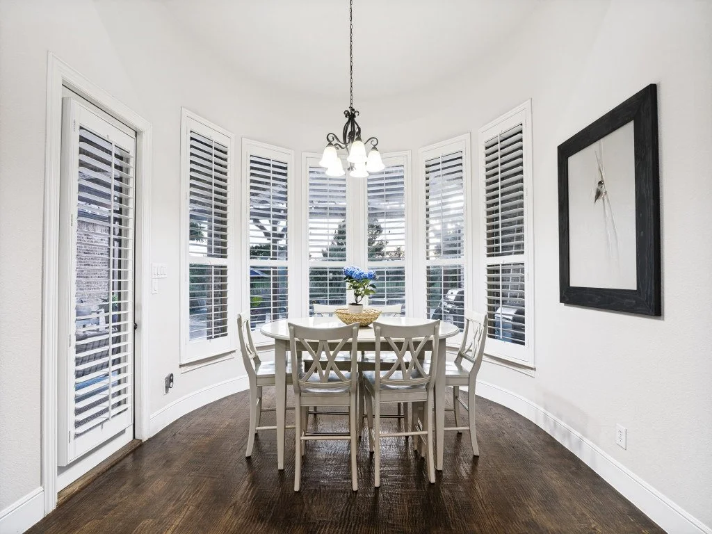 A dining area with a round table, six chairs, a chandelier, a flower vase with blue flowers, a window with white shutters, and a framed artwork on the wall.