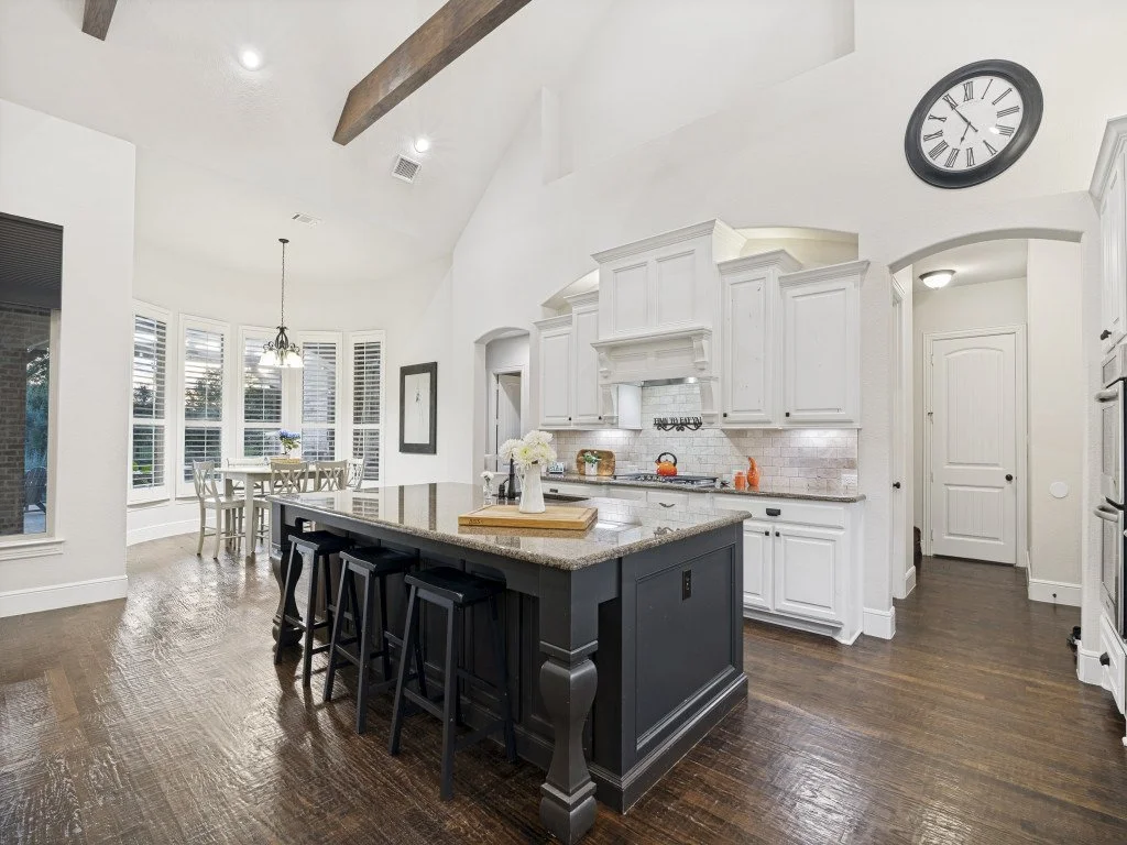 Modern kitchen with white cabinets, black island, hardwood floors, dining area with large bay window, and wall clock.