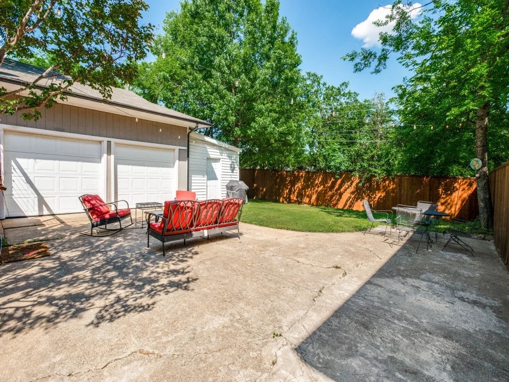 Backyard patio with outdoor furniture, including a red cushioned loveseat, chairs, and a small table. A grassy area with trees and a wooden fence surrounds the space on a sunny day.