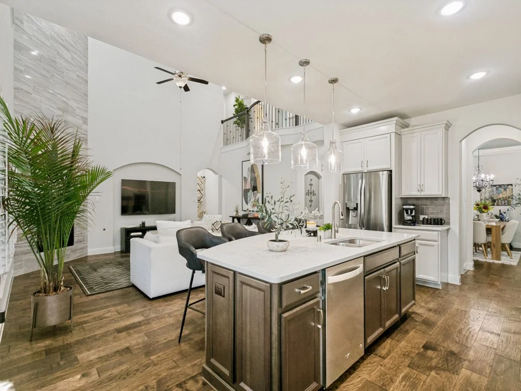 Open-concept kitchen and living room with modern decor, white cabinets, stainless steel appliances, marble island, pendant lights, dining area, and large potted plant.