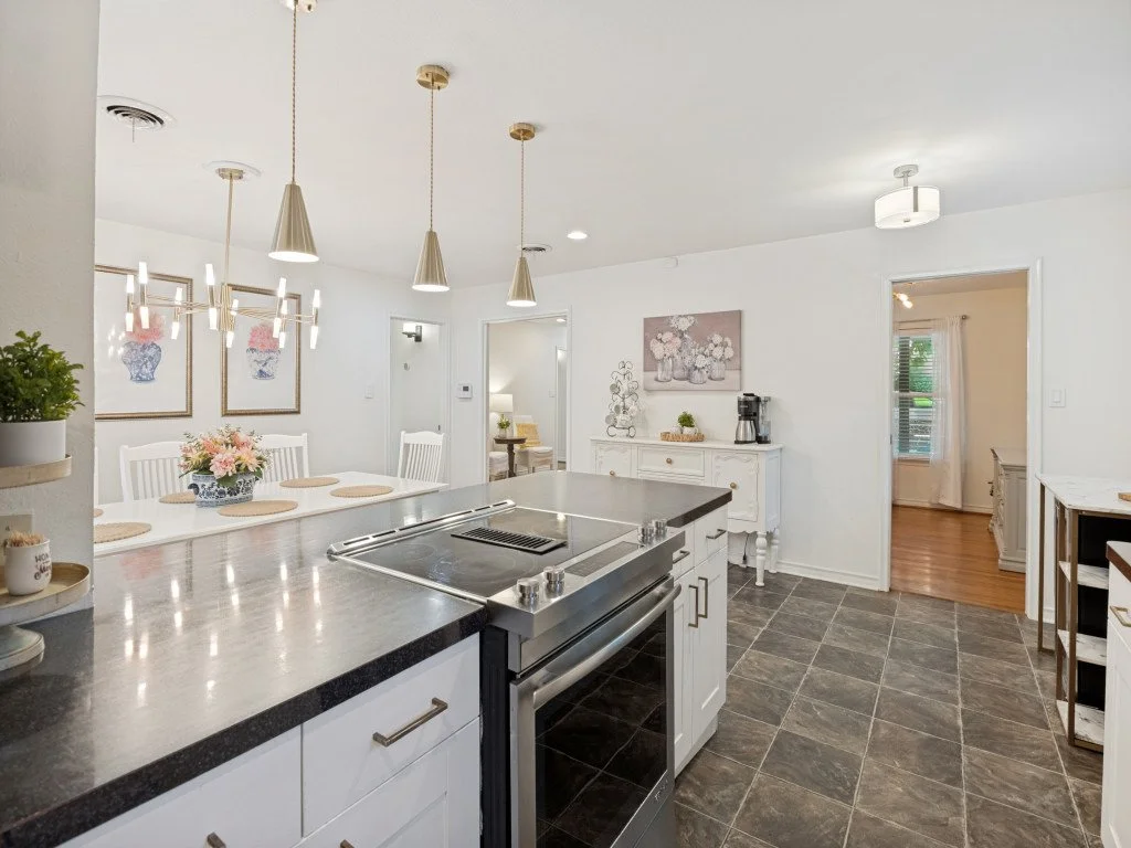 Bright, modern kitchen with white cabinetry, black countertop, stainless steel oven, and decorative lighting fixtures. Adjacent dining area with white table and chairs, floral centerpiece, and wall art.