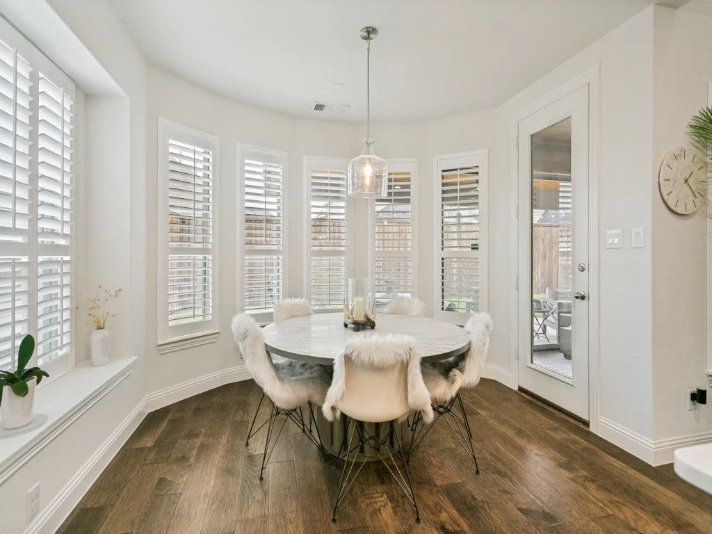A dining room with a round table, six chairs with white fluffy covers, and a glass pendant light, surrounded by large windows with white plantation shutters.