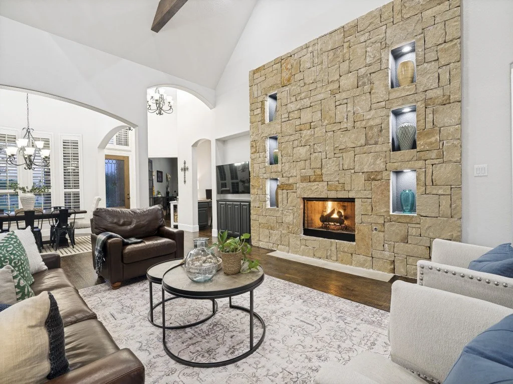 Cozy living room with a stone fireplace, modern built-in display niches, decorated with vases and small sculptures, with sofas, a coffee table, and a rug, adjacent to a dining area with a chandelier and large window with white shutters.