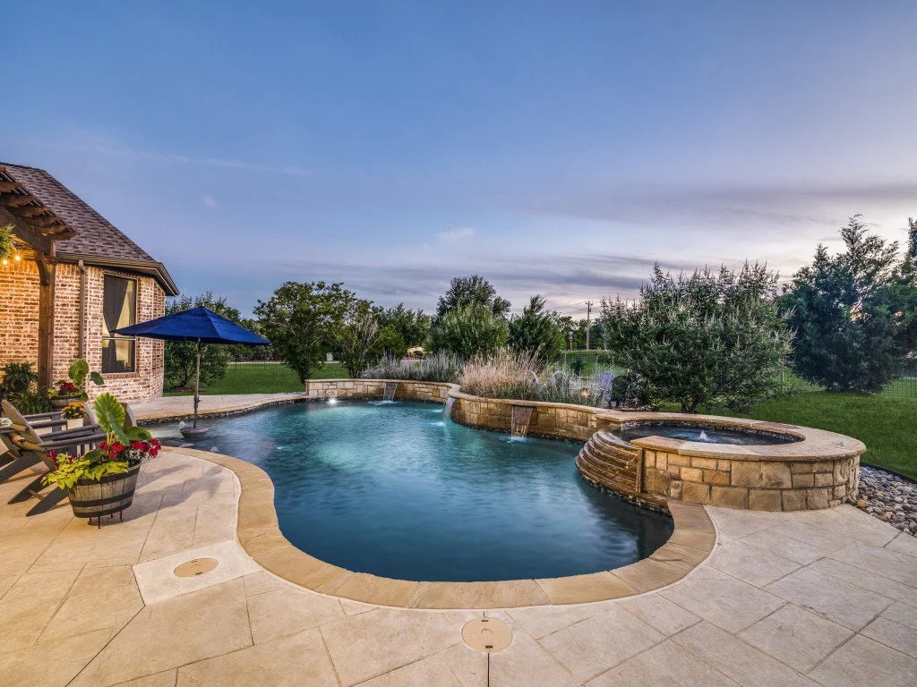 Backyard with a swimming pool, hot tub, and stone patio. A house with brick exterior and window on the left, and trees and grass in the background, during dusk.