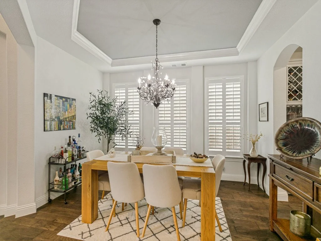 A dining room with a wooden table, six beige upholstered chairs, a chandelier, three windows with plantation shutters, a painting on the wall, a bar cart with bottles, and a side table with a white vase.