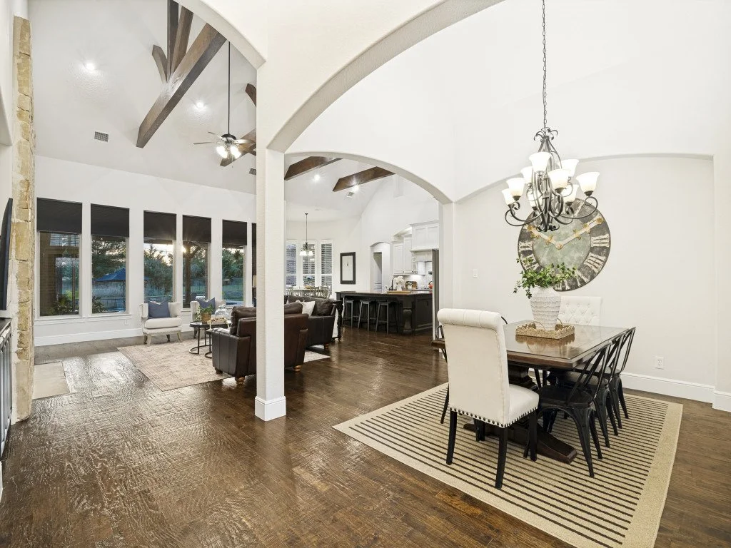 Open-concept living and dining area with hardwood floors, white walls, large windows with black shades, a stone fireplace, and a dining table with white and black chairs, a chandelier, and a large wall clock.