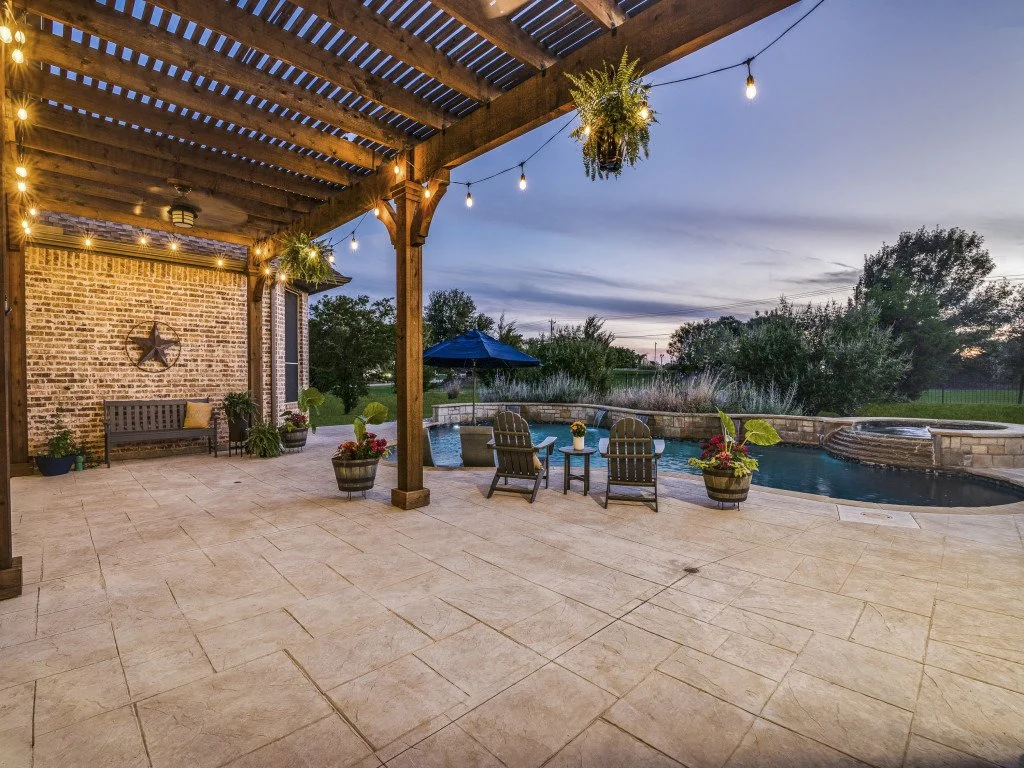 Outdoor patio with string lights, hanging plants, potted plants, a brick wall with a metal star, a bench, Adirondack chairs around a small table, a swimming pool, and trees in the background during dusk.