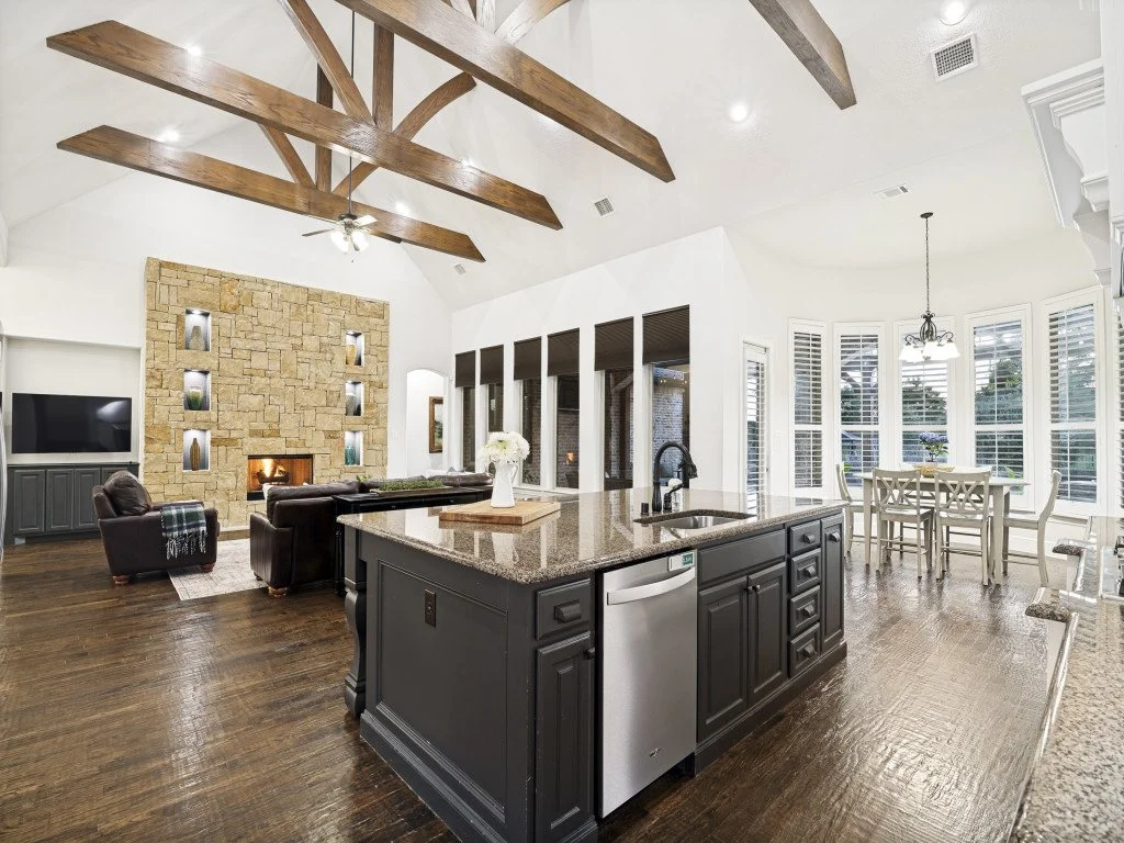 Open-concept living room and kitchen with vaulted ceiling, exposed wooden beams, stone fireplace, dark wood flooring, and large windows with white shutters.