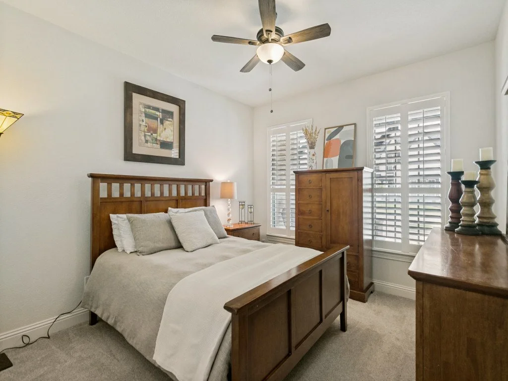 A bedroom with a wooden bed frame, white bedding, gray pillows, a nightstand with a lamp, a wooden dresser, and a tall wooden chest, with windows covered by white shutters and a ceiling fan.