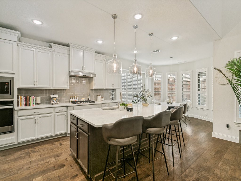Modern kitchen with white cabinets, a central island with a white countertop, gray bar stools, pendant lights, multiple windows with shutters, and hardwood floors.