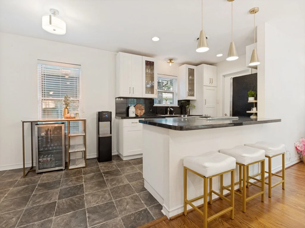 Kitchen with white cabinets, black countertop, pendant lights, and bar stools with white cushions and gold legs.