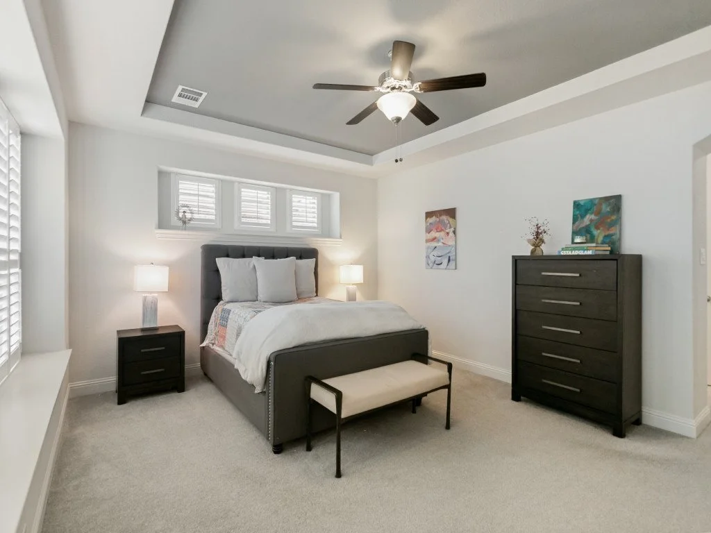 A bedroom with a large bed, dark gray headboard, white bedding, two nightstands with lamps, a bench at the foot of the bed, a tall dresser with artwork and a decorative vase on top, a ceiling fan with a light, white walls, a window with plantation sh