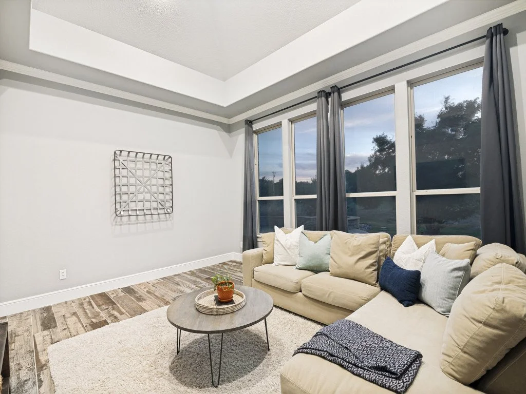Living room with beige sectional sofa, light-colored pillows, a round gray coffee table with a plant, dark curtains, large windows showing trees outside, and a white rug, in a modern home interior.