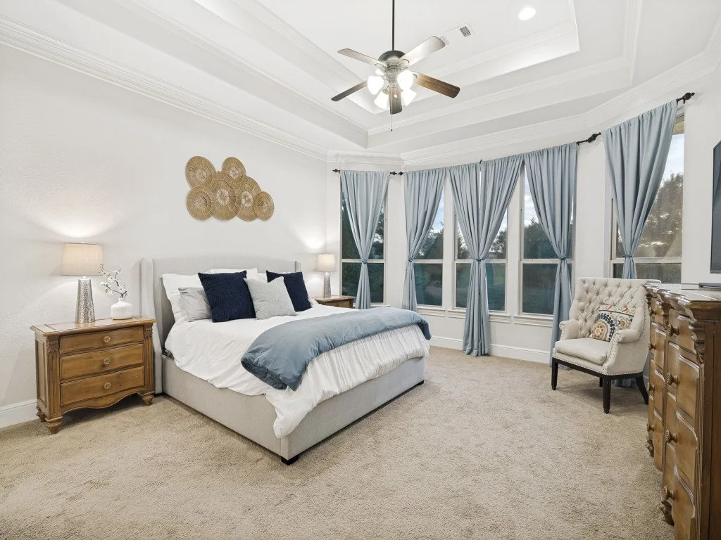 A bedroom with a large bed, nightstands, lamps, and a tufted armchair. The room has multiple windows with light blue curtains, a ceiling fan, and beige carpet.