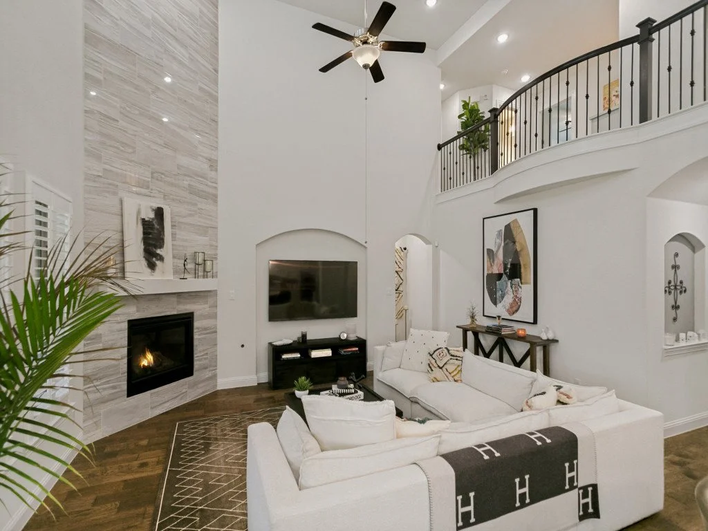 Living room with white sectional sofa, dark wood floor, fireplace, flat-screen TV, abstract wall art, ceiling fan, upstairs balcony, decorative pillows, and houseplants.