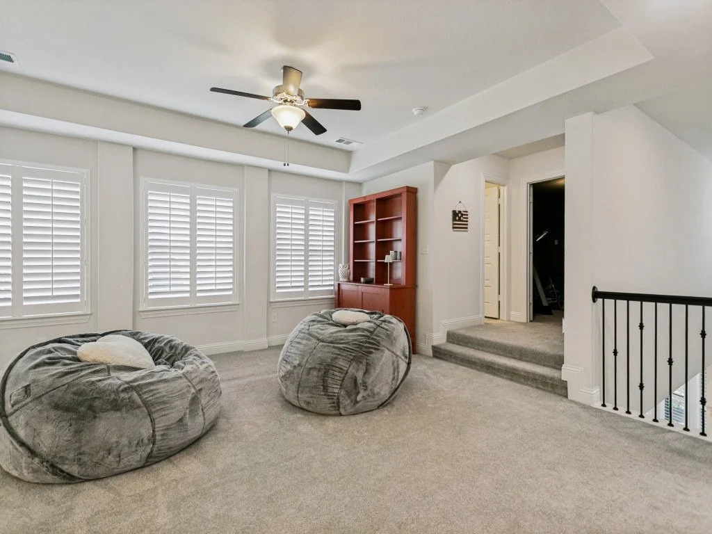 Living room with three large windows with white shutters, beige carpet, two gray bean bag chairs, a ceiling fan, a wooden bookshelf, and an open doorway leading to a stairway and hallway.