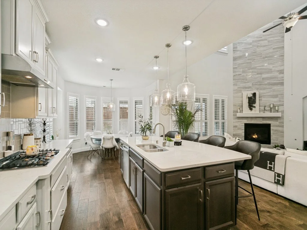 Modern open-concept kitchen with white cabinets, a central island with dark lower cabinets, pendant lights, and a living area with a fireplace and bay windows.