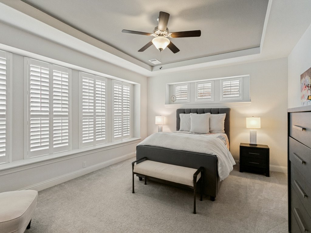A bedroom with white walls, a gray upholstered bed, and white bedding. There are two nightstands with lamps, a window with blinds above the bed, a ceiling fan, and a gray dresser.