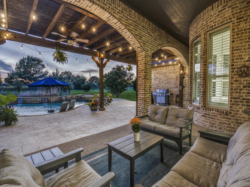 Outdoor covered patio with brick arches, string lights, and patio furniture overlooking a pool and a yard with trees and a blue umbrella.