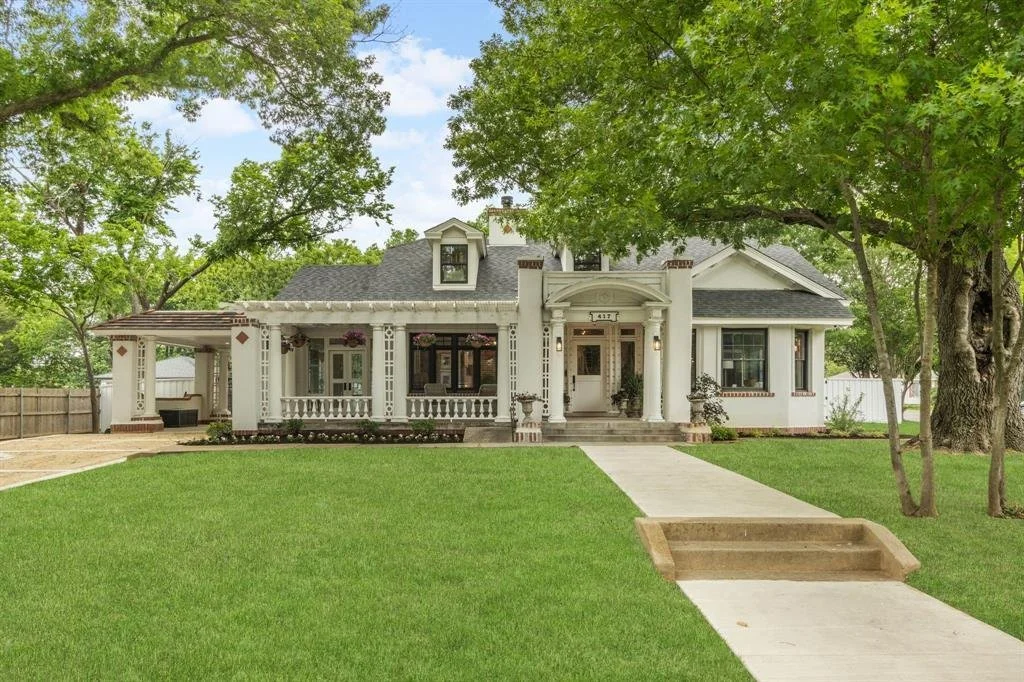 A large white house with a front porch, surrounded by green trees and a well-kept lawn, in a suburban neighborhood.
