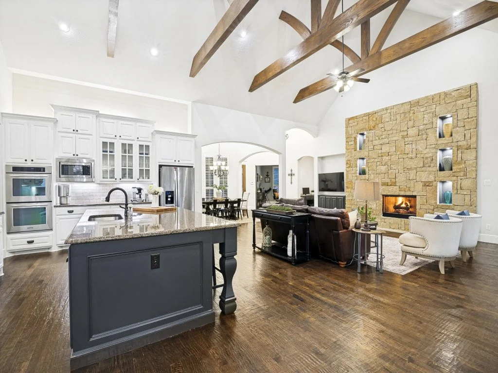 Open-concept living room and kitchen with white cabinetry, stainless steel appliances, a stone fireplace, wood beams on the ceiling, and hardwood floors.