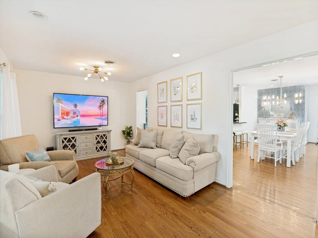 Living room with white walls, hardwood floors, a beige sofa, beige armchair, a glass coffee table, and a TV on a gray console. Art frames hang on the wall, and a doorway leads to a dining room with white furniture and patterned wallpaper.