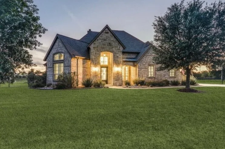 Large stone and brick house with illuminated windows, surrounded by a well-maintained lawn and trees at dusk.