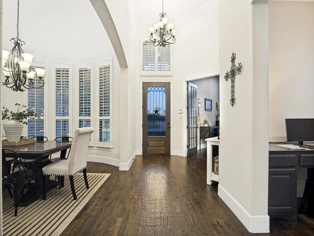 Interior view of a home entry hall with dark hardwood floors, a wooden door with a window, and a chandelier overhead. There is a dining area to the left with a table, chairs, and window shutters, and a small desk with a computer to the right.