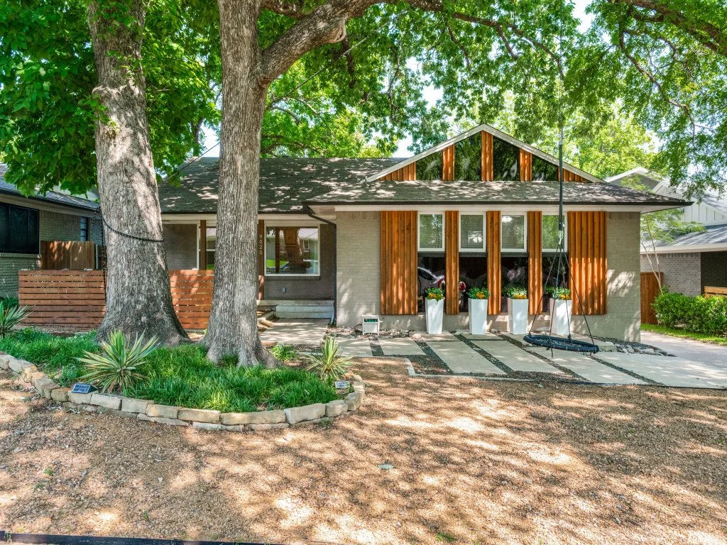 A modern house with large windows, wooden accents, and a front porch, surrounded by trees and some landscaping, including potted plants and a swing.