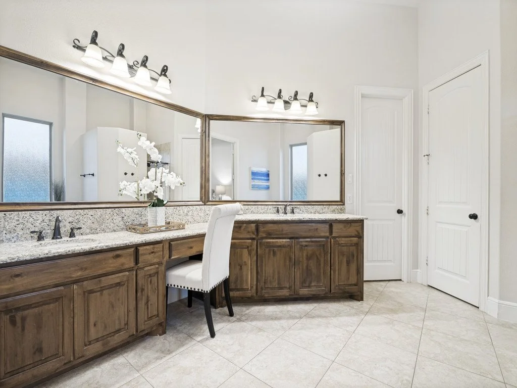 A bathroom with a long wooden vanity, granite countertop, large mirror, two light fixtures, a white upholstered chair, and white walls.