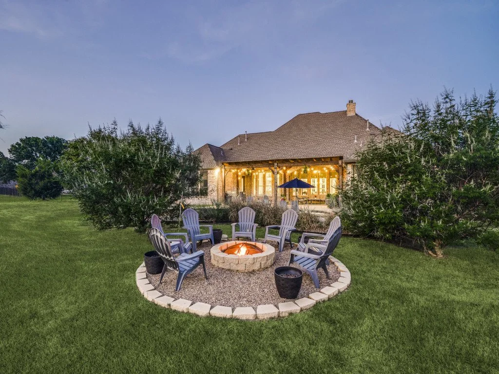 Backyard with lit fire pit surrounded by six Adirondack chairs, bushes, and a house with lit windows in the background during dusk.