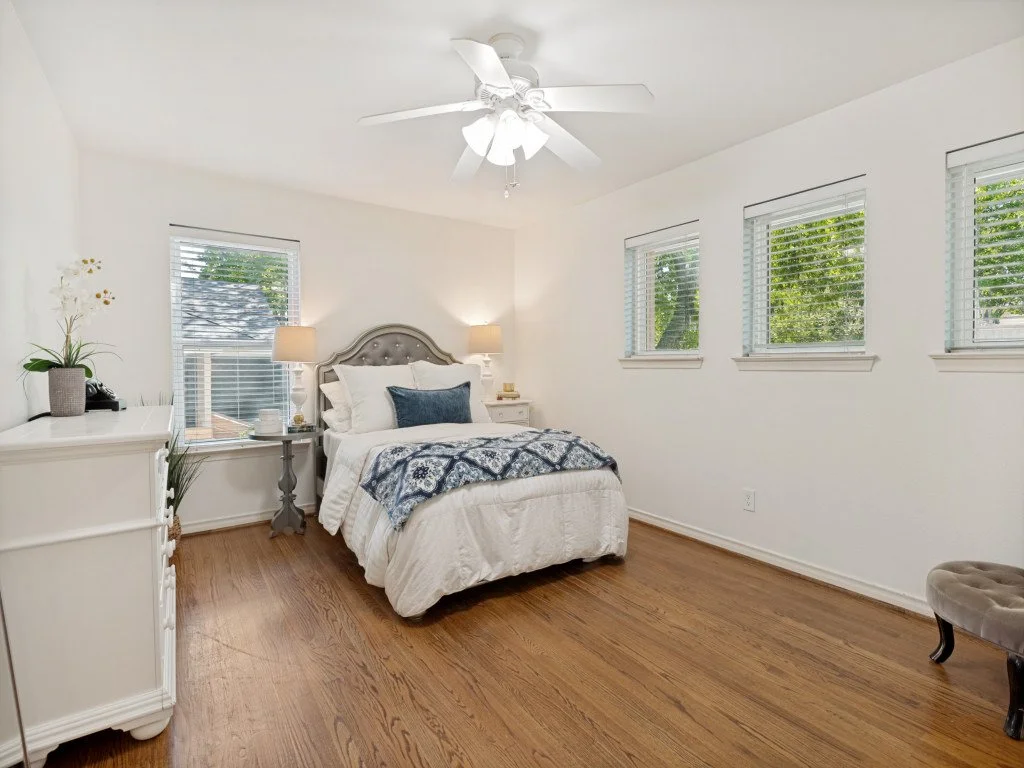 A bedroom with white walls, hardwood floor, and a ceiling fan. It features a bed with white bedding and a blue throw blanket, two white nightstands with lamps, and three windows with white blinds letting in natural light.