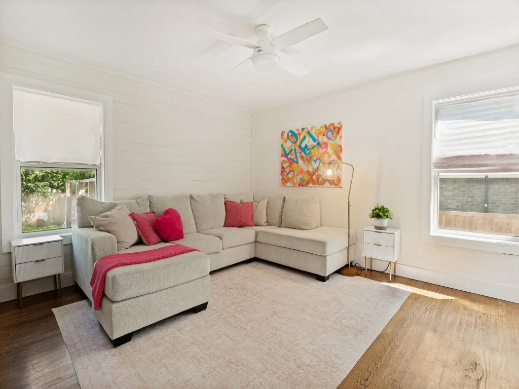 Bright living room with white walls, a beige sectional sofa with pink and red pillows, a white side table, a potted plant, colorful artwork on the wall, a floor lamp, and hardwood floors.