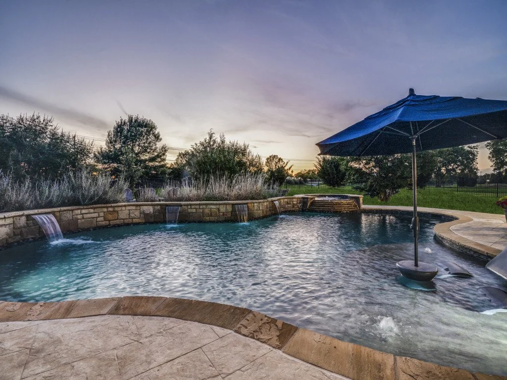 A backyard swimming pool with a stone and brick water feature and a large blue umbrella at sunset.