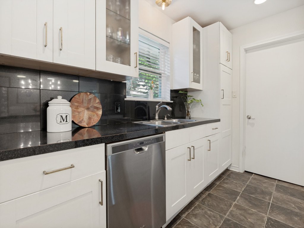 Kitchen with white cabinets, black countertop, gray tiled floor, and a window above the sink. On the counter, there is a white container with an 'M' monogram, a cutting board, and a small plant.