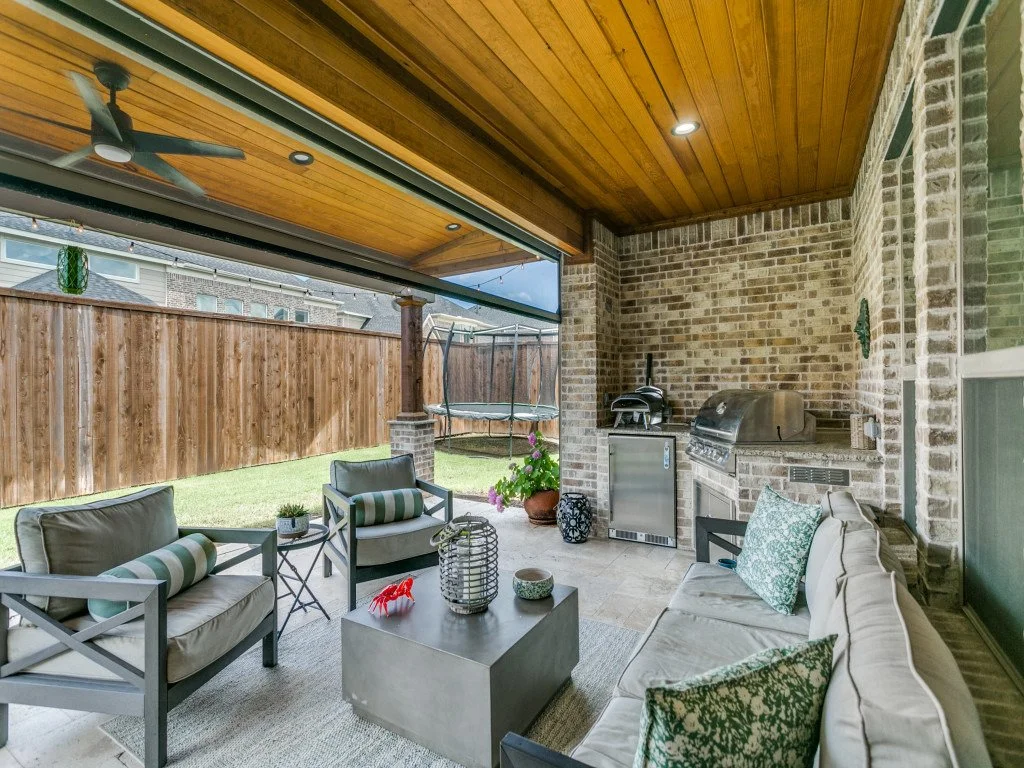 Covered outdoor patio with seating area, ceiling fan, brick wall, grill, mini fridge, and trampoline in backyard.