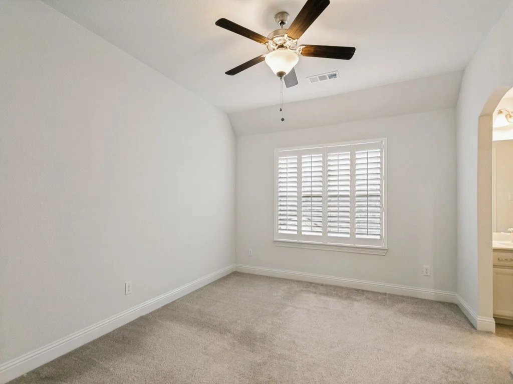 Empty bedroom with white walls, window with white shutters, beige carpet, ceiling fan with lights, and an arched doorway to another room.