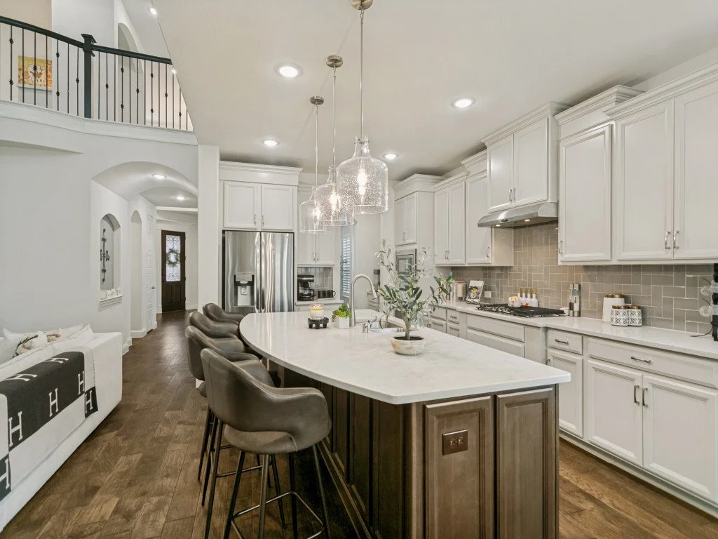 Modern kitchen with white cabinetry, gray backsplash, and a large island with a marble countertop. Hanging pendant lights above the island and stainless steel appliances.