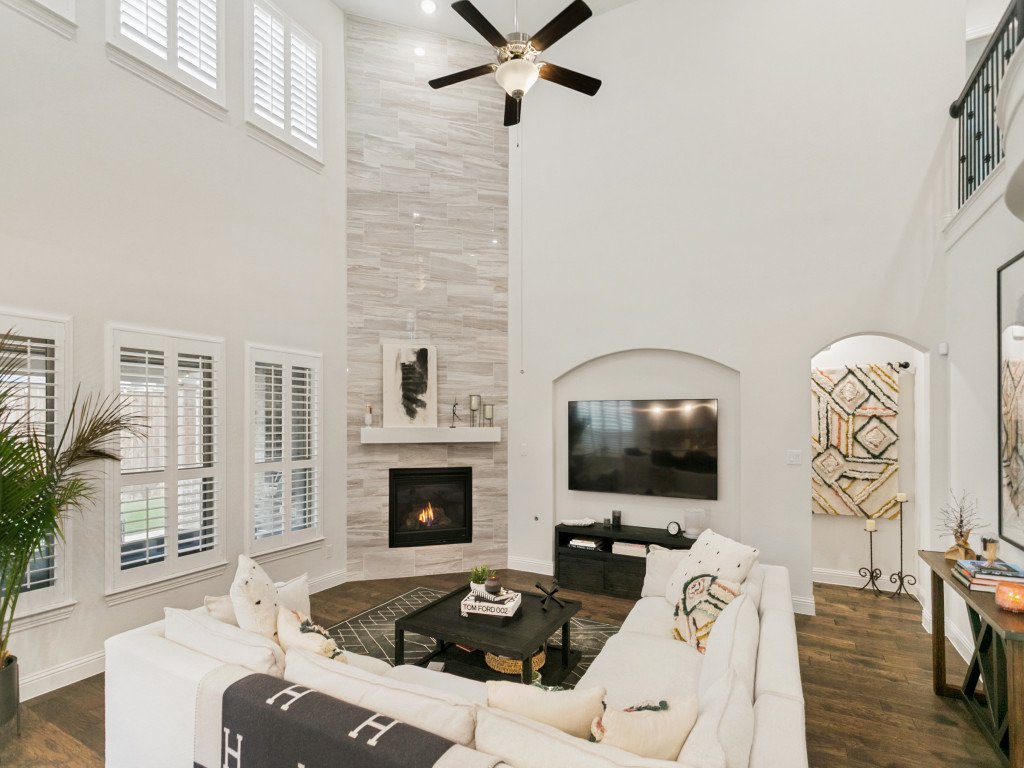 Modern living room with high ceiling, large windows with white plantation shutters, a wall-mounted TV, a fireplace with a tall gray tile accent wall, a black ceiling fan, and white sofas with decorative pillows. The room features dark wood flooring, 